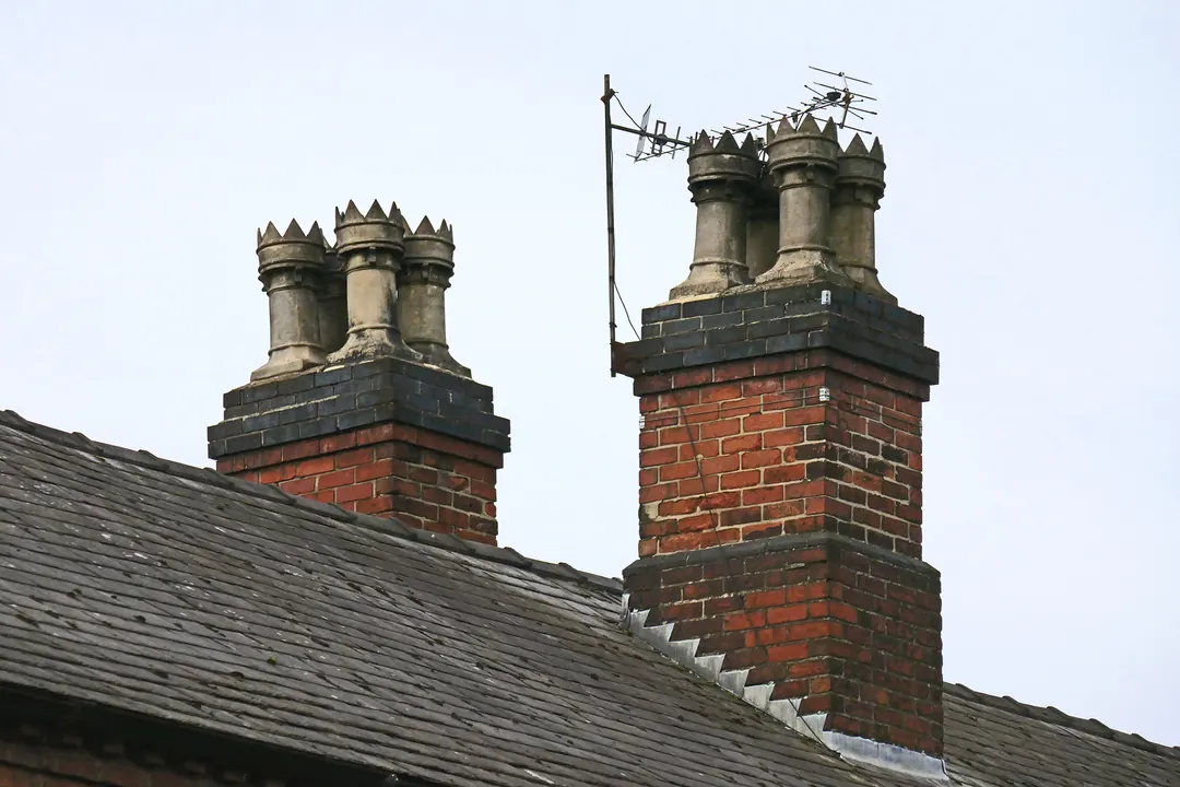 Two red brick chimney stacks with clay pots rising from a weathered slate roof against a pale grey sky