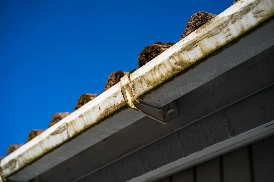 Stained white plastic gutter with a jointing bracket, running beneath mossy concrete roof tiles
