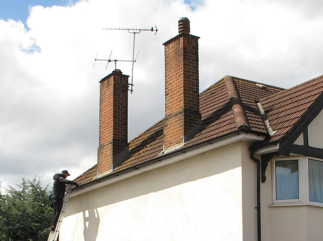 Roofer on a ladder at the eaves checking the gutter and tiled roof of a house with two brick chimney stacks