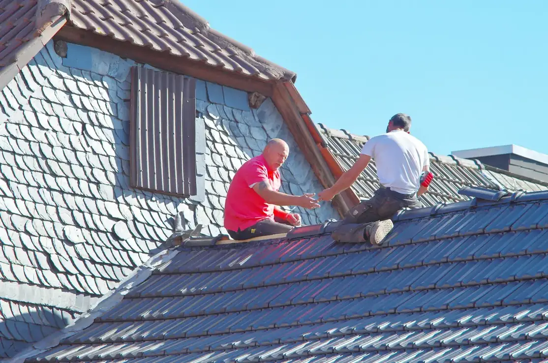 Two roofers kneeling on a slate roof beside a tile-hung dormer, working on a repair